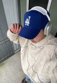 Casquette de baseball bleue avec logo blanc, pull crème en tricot torsadé et écouteurs argentés. Le sujet se tient sur une surface en béton près d'un garde-corps.