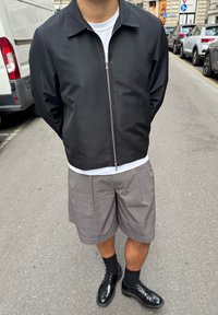 Crna, lagana jakna na patentni zatvarač s ovratnikom; uparena sa sivim kariranim šortsevima; crnim cipelama; čarape do koljena u crnoj boji.