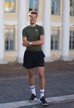 Camiseta de compresión verde, pantalones cortos negros con logo, calcetas blancas a rayas y zapatillas negras. De pie al aire libre, postura relajada, fondo neutral.