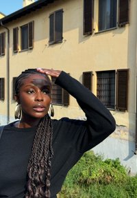 Maglione nero con capelli lunghi e intrecciati, grandi orecchini a cerchio dorati e una mano alzata sulla fronte, con un edificio giallo sfocato sullo sfondo.