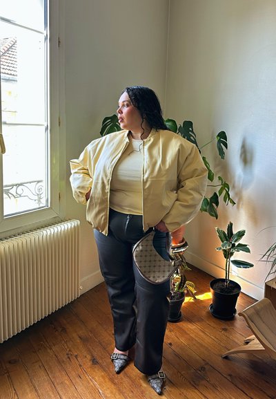 Mujer con chaqueta amarilla y pantalones negros está junto a una ventana en una habitación con suelos de madera y plantas de interior en macetas, sosteniendo un pequeño bolso.