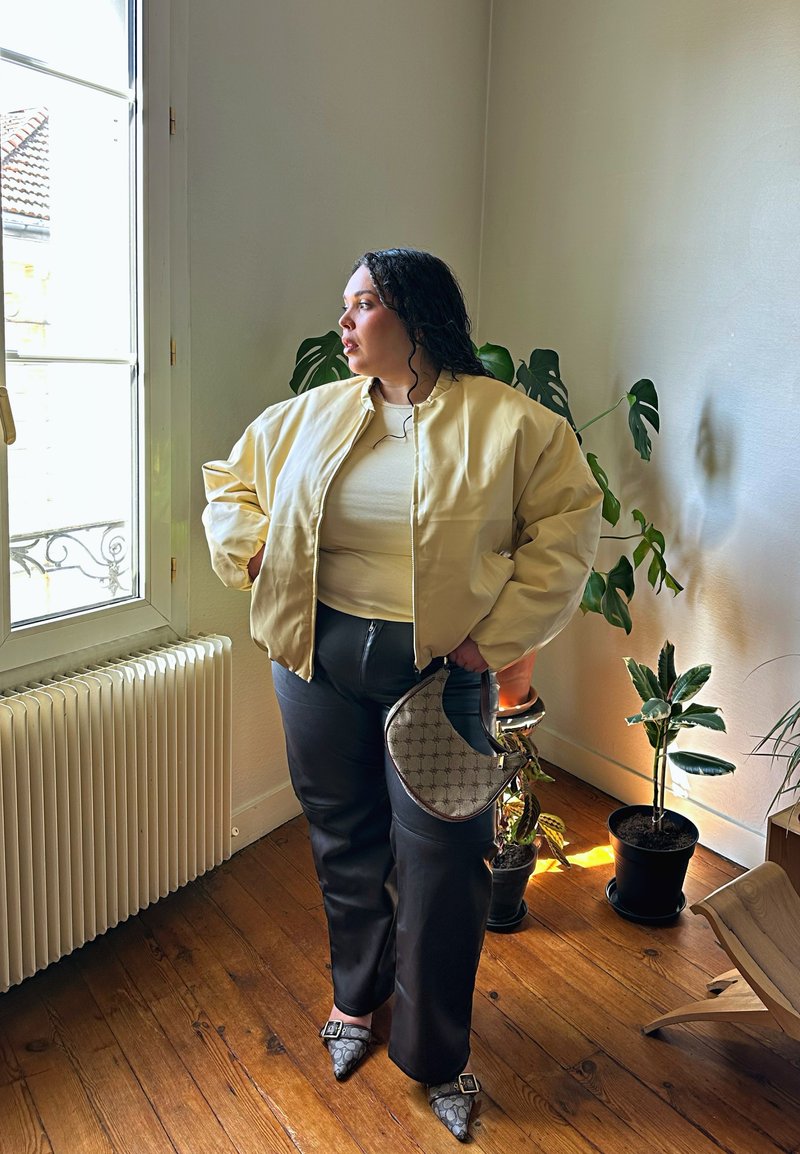 Γυναίκα με κίτρινο μπουφάν και μαύρο παντελόνι στέκεται δίπλα σε ένα παράθυρο σε ένα δωμάτιο με ξύλινο πάτωμα και εσωτερικά φυτά σε γλάστρες, κρατώντας μια μικρή τσάντα.