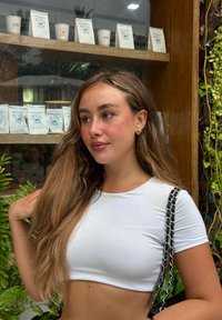 Jeune femme aux longs cheveux châtain clair portant un crop top blanc et des boucles d'oreilles dorées, se tenant près de verdure et d'une étagère en bois avec des emballages de café.