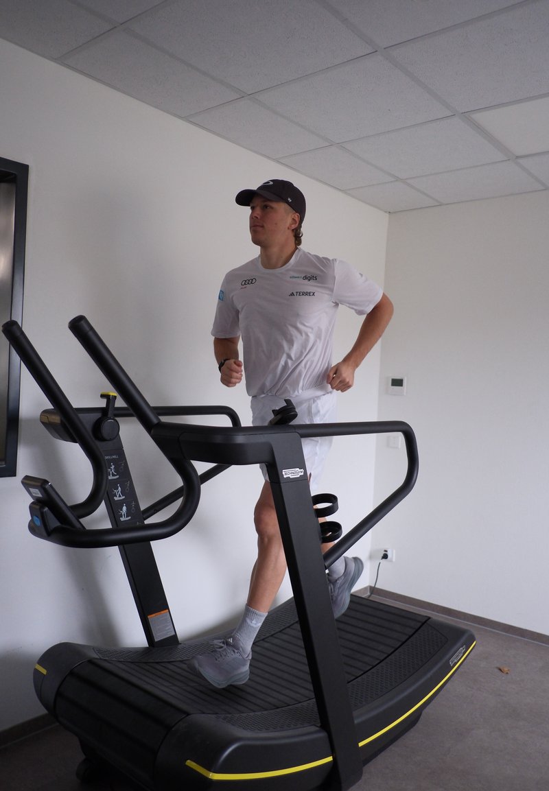 Tapis roulant con struttura nera e superficie di corsa testurizzata, con accenti gialli. Una persona in abbigliamento bianco sta facendo jogging.