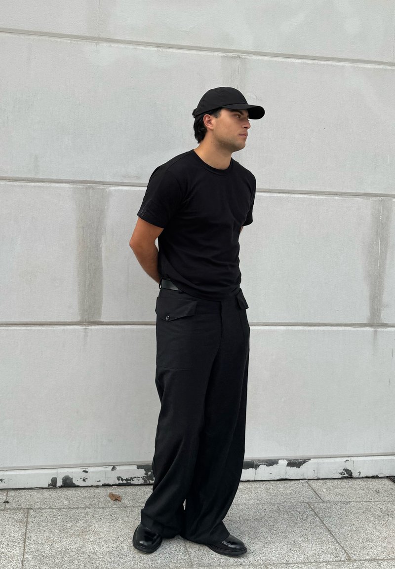 T-shirt noir à manches courtes, pantalon noir ample avec poches latérales, et casquette noire, se tenant contre un mur gris clair.