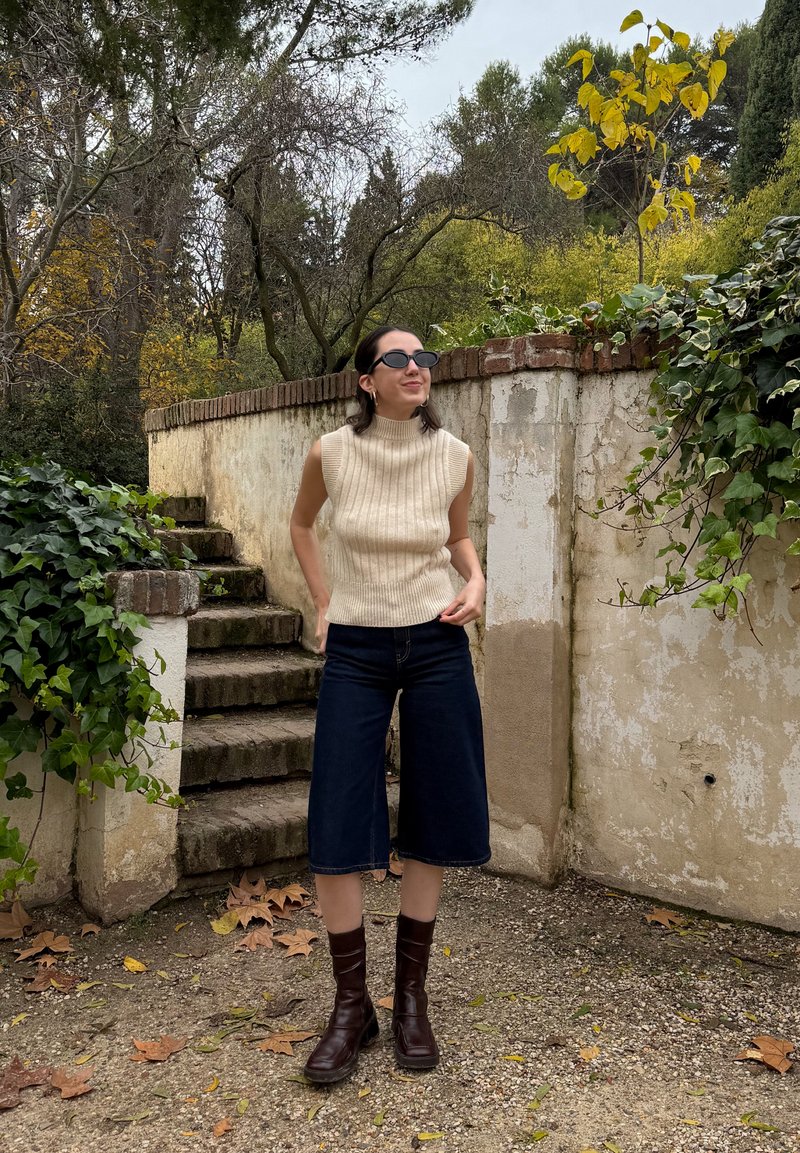 Μπεζ ριγέ αμάνικο λαιμόκοψη pullover, σκούρα τζιν κολάν, και καφέ μπότες μέχρι το γόνατο, σε φόντο με υφή από φυτά και πέτρα.