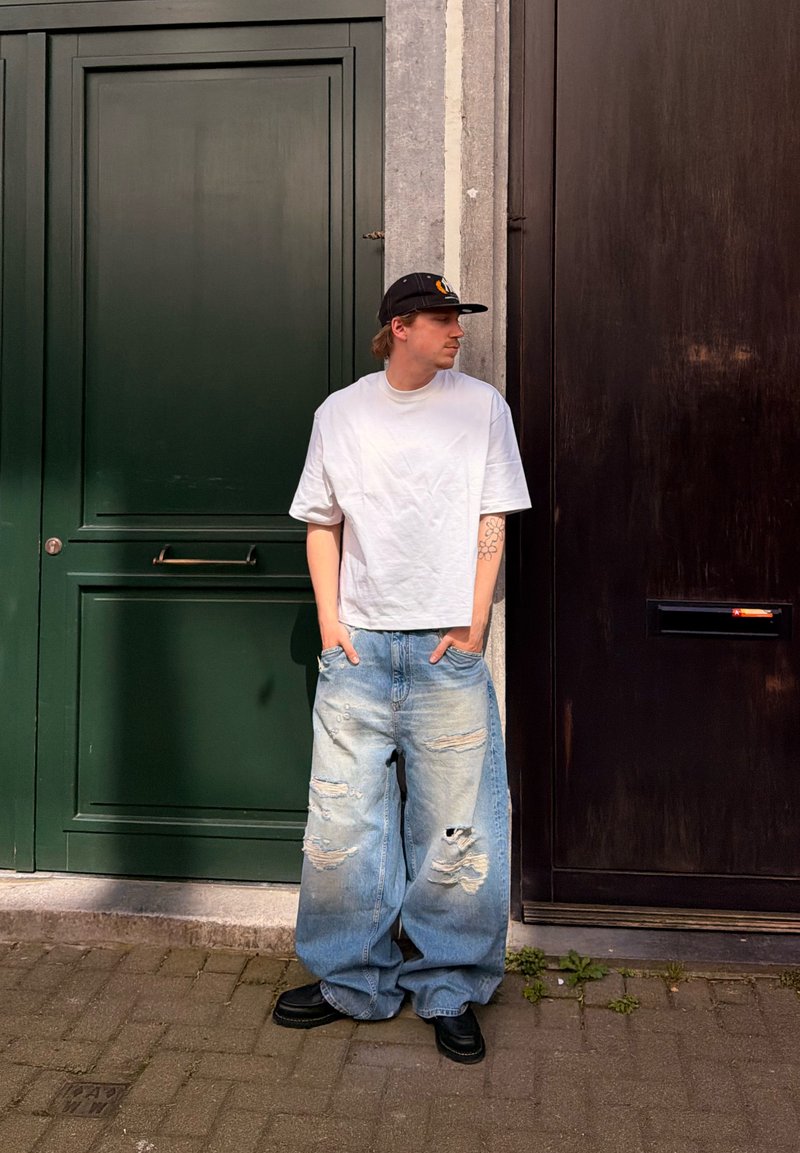 Homme portant un t-shirt blanc ample, un jean déchiré et large, une casquette noire et des chaussures, debout les mains dans les poches entre des portes vert foncé et marron foncé.