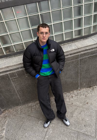 Chaqueta negra acolchada, suéter verde con rayas azules y negras, vaqueros oscuros y zapatos plateados. Gafas delgadas y cabello corto. De pie contra una pared.