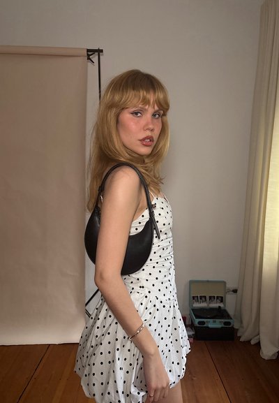 Bolso de hombro de cuero negro con forma semicírculo. El modelo lleva un vestido blanco con puntos negros. Textura suave, minimalismo en los accesorios.