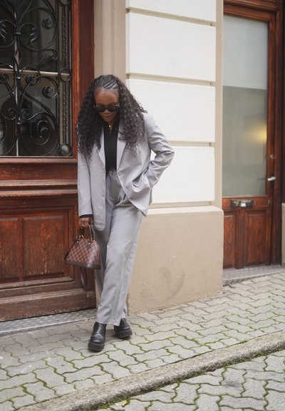 Mujer con cabello largo y rizado, vestida con un traje gris claro, botas negras y gafas de sol, caminando por una calle empedrada con un bolso marrón de cuadros en la mano.