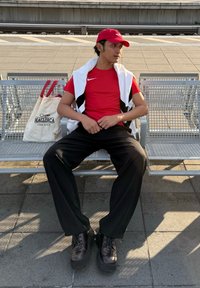 Maglietta rossa Nike, pantaloni neri e scarpe platform marroni. Asciugamano bianco sulle spalle. Borsa beige con la scritta "MALLORCA ONE LOVE" accanto a lui.