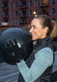 Fekete gyógylabda fekete kesztyűben tartva, texturált felülettel. A személy világoskék hosszú ujjú felsőt és fekete mellényt visel.