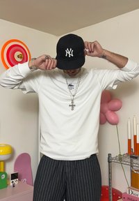 Witte langemouwen shirt met Lacoste-logo, zwarte New York Yankees-pet, zilveren kettingen en gestreepte broek. Ronde en gestreepte wanddecoratie.