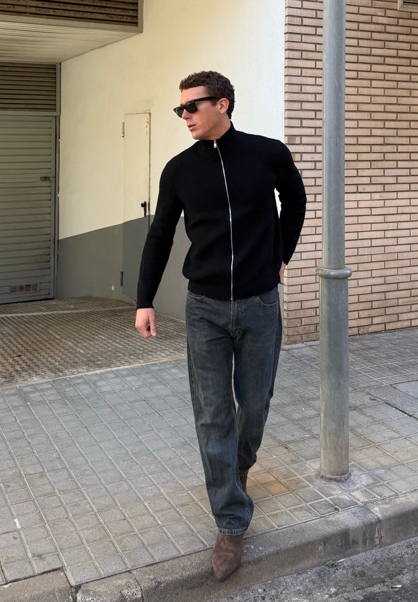 Muž v čiernom svetri na zips, tmavých džínsoch a hnedých topánkach kráčajúci po mestskom chodníku, má na sebe slnečné okuliare a pozerá do strany.