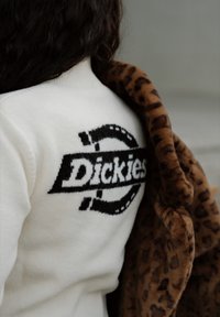 Valge fleece-jope musta "Dickies" logoga. Prantsuse leopardikujuline vale-sisuga wrapped on hoitud ühe õla peal.