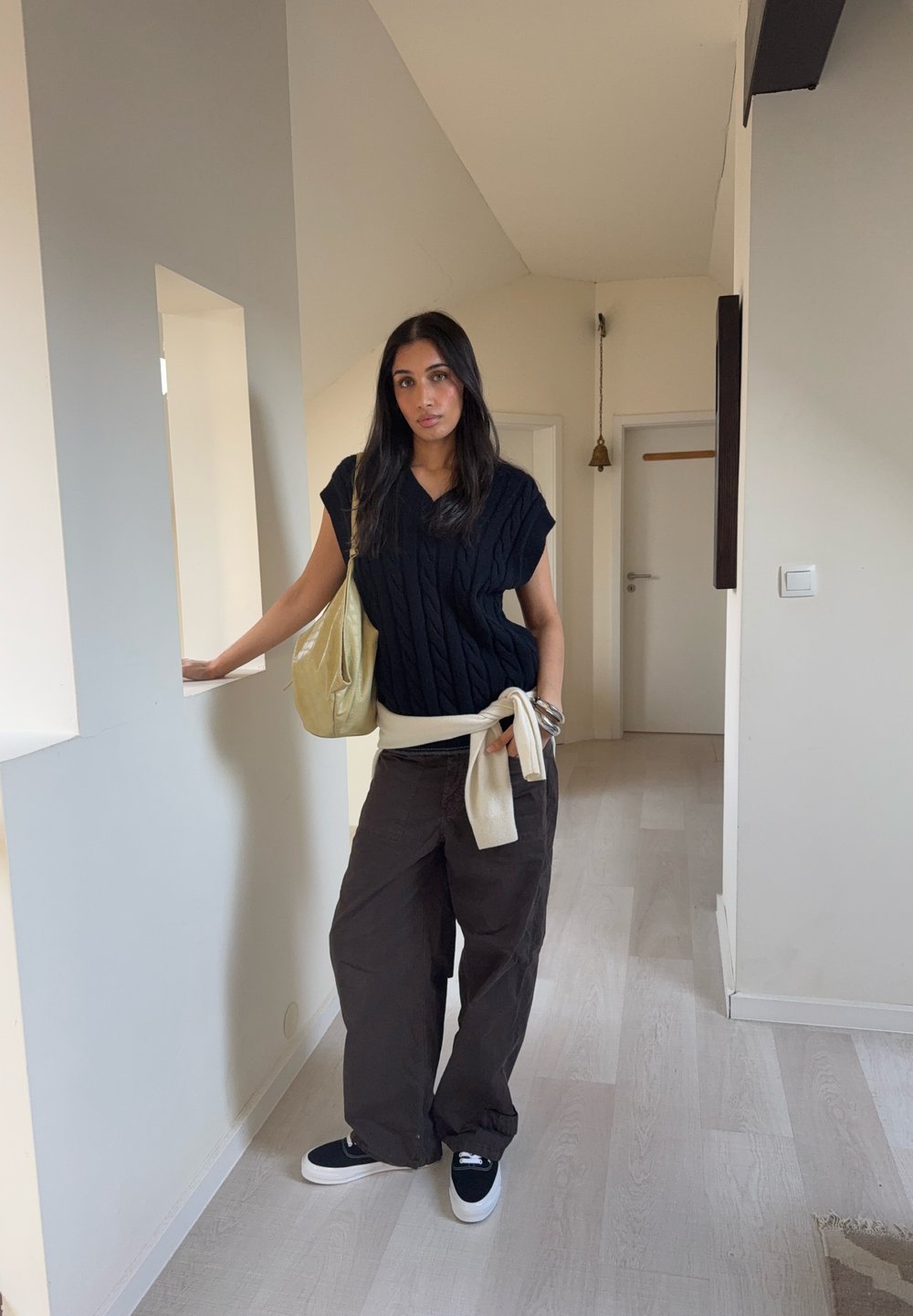 Haut noir en maille torsadée, ceinture à la taille beige clair, pantalon large marron, baskets noires avec semelles blanches, et sac à bandoulière beige clair.