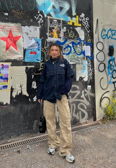 Camisa de mezclilla de botones sobre una camiseta de manga larga, pantalones cargo beige y zapatillas gruesas. Bolso negro con tachuelas. Fondo de pared cubierto de graffiti.