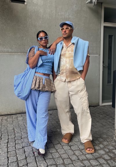 Dos personas posan frente a una pared gris texturizada. Una lleva una blusa de rayas azules y pantalones, la otra una camisa beige con estampado y pantalones de carga.