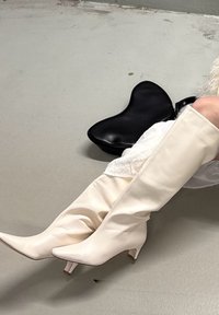 Botas de couro brancas até ao joelho com bico pontiagudo e salto baixo, acompanhadas por uma mala preta em forma de coração. Textura suave.