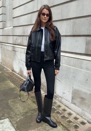 Mujer con chaqueta de cuero negra, pantalones negros y botas altas negras sosteniendo un bolso negro, de pie junto a una pared de piedra de un edificio.