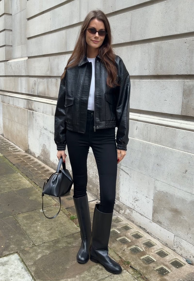 Femme en veste en cuir noire, pantalon noir et bottes hautes noires tenant un sac à main noir, debout à côté d'un mur de bâtiment en pierre.