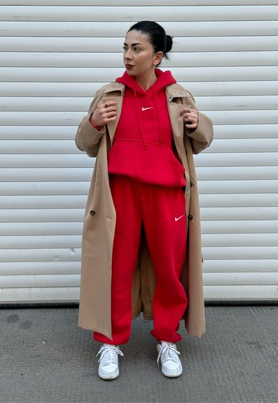 Sudadera roja de Nike con pantalones jogger a juego, todo bajo un abrigo trench beige. Zapatillas blancas completan el look. El fondo es una puerta de garaje blanca.