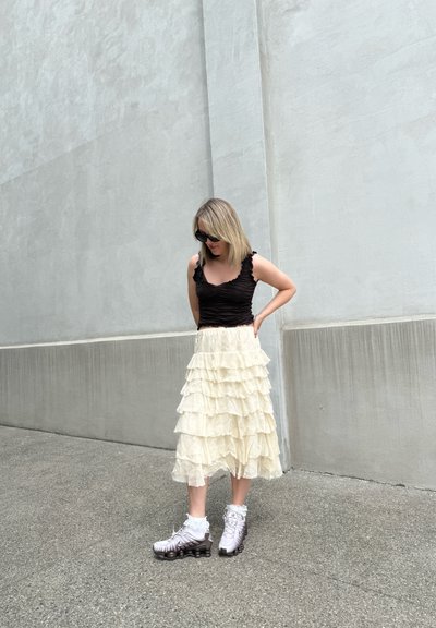 Top corto negro fruncido combinado con una falda en capas crema, que cuenta con volantes. El calzado incluye zapatillas deportivas blancas con detalles oscuros.