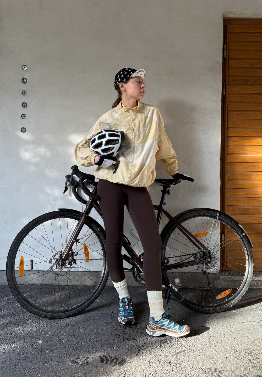 Light yellow windbreaker with white stripes, brown leggings, black and white cycling helmet, blue and brown trainers, bicycle with a black frame.