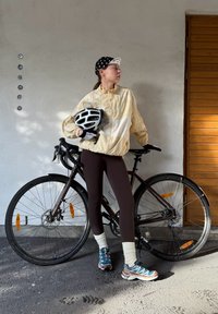 Light yellow windbreaker with white stripes, brown leggings, black and white cycling helmet, blue and brown trainers, bicycle with a black frame.