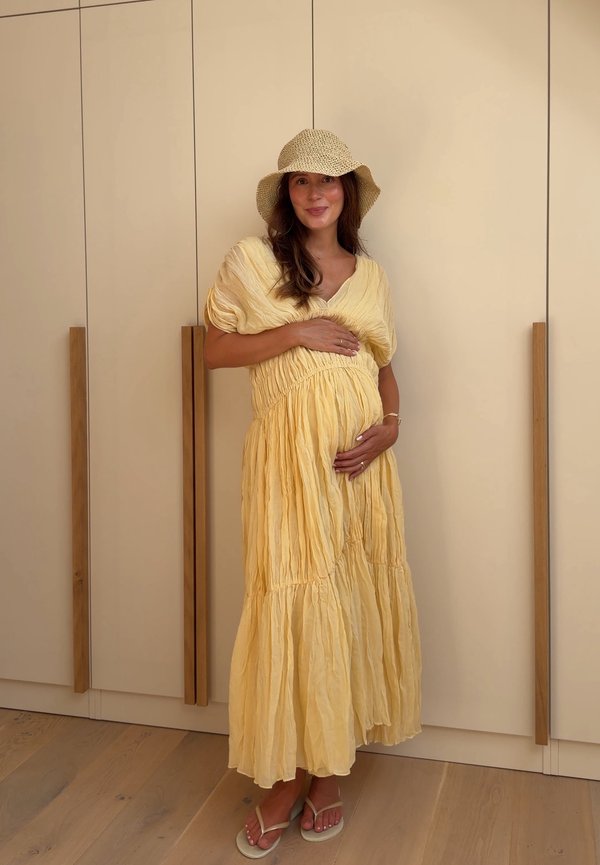 Femme enceinte en robe jaune et chapeau tressé debout pieds nus sur un sol en bois, tenant son ventre et souriant devant une armoire beige.