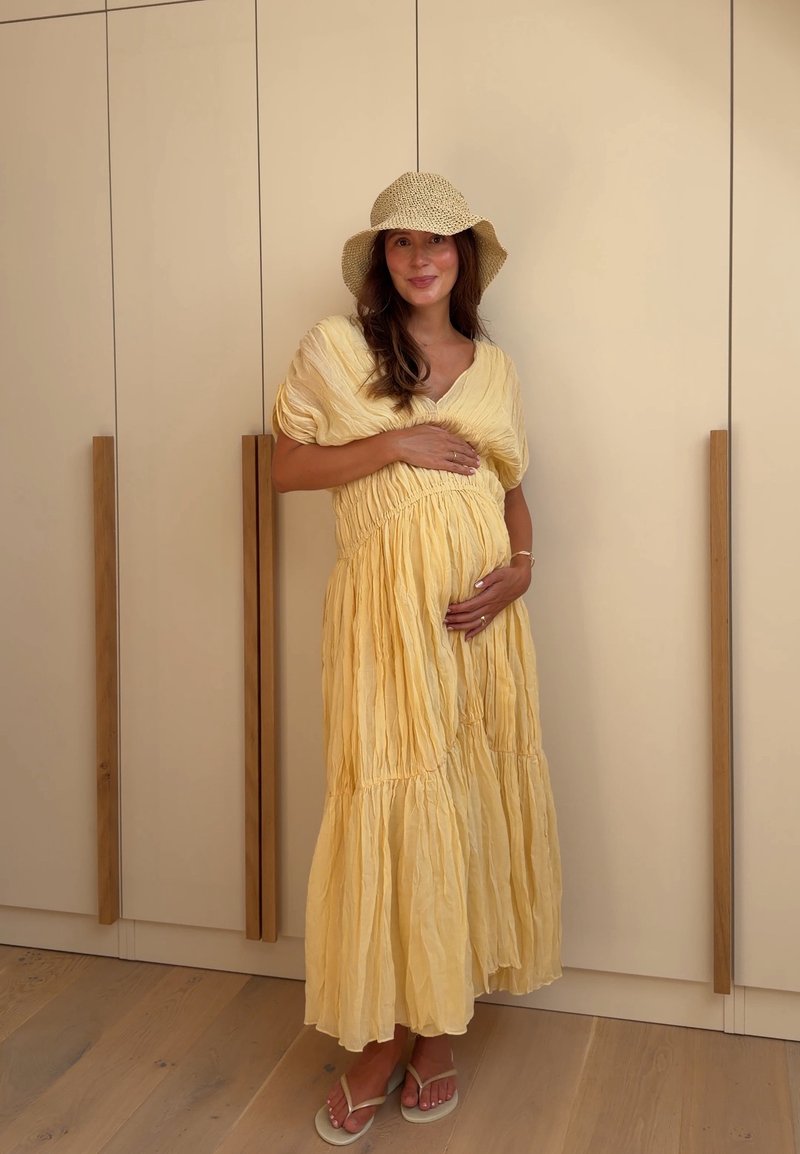 Femme enceinte en robe jaune et chapeau tressé debout pieds nus sur un sol en bois, tenant son ventre et souriant devant une armoire beige.