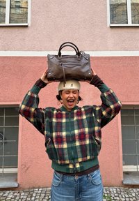 Jeune femme portant une veste à carreaux et un chapeau blanc, tenant un sac en cuir marron au-dessus de sa tête, debout devant un mur rose et rouge avec des fenêtres.