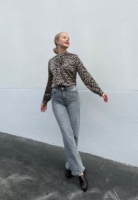 Μπλούζα με λεοπάρ σχέδιο και λαιμόκοψη που δένει, συνδυασμένη με ανοιχτό γκρι φαρδιά τζιν και σκούρους loafers, σε φόντο απλού λευκού τοίχου.