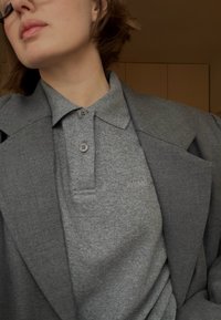 Polo gris avec une finition texturée et une patte de boutonnage à trois boutons, porté sous un blazer gris assorti, avec une broderie de logo discrète.