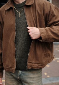 Jachetă din corduroi maro descheiată peste un pulover verde închis, cu o textură netedă și un colier din lanț auriu vizibil. Se asortează cu blugi albaștri deschiși.