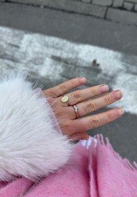 Anello sigillo in oro con dettagli incisi, accompagnato da un anello in oro rosa con tre piccole pietre, posati su una sciarpa rosa e pelliccia bianca.