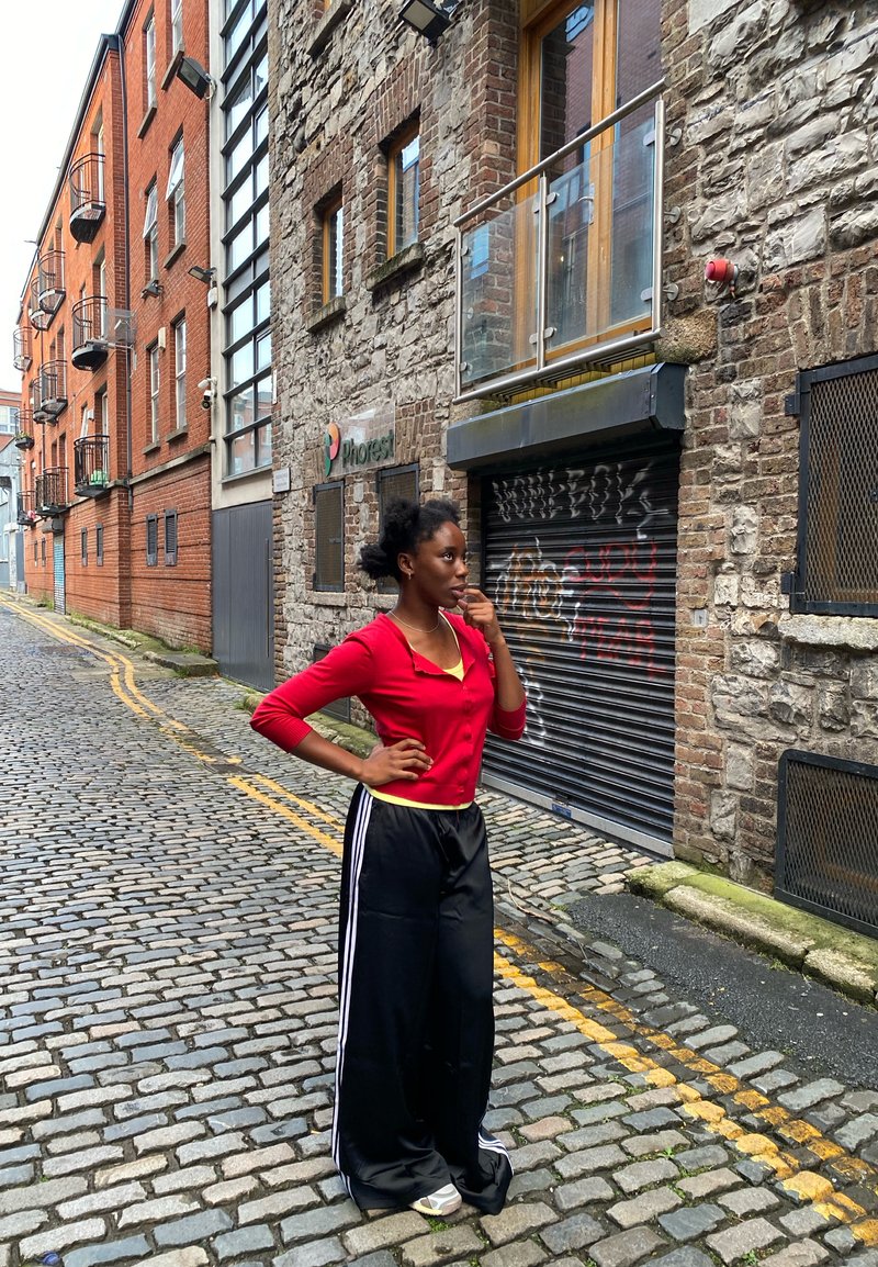 Cardigan roșu mulat, bluză galbenă de sub, pantaloni negri evazați cu dungi albe, stând pe o stradă pietruită lângă clădiri din cărămidă.