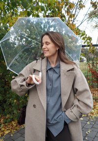 Um guarda-chuva transparente segurado por uma pessoa vestindo um casaco bege sobre uma camisa listrada azul. O fundo apresenta folhagem verde e folhas de outono.
