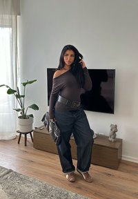 Haut marron à épaules dénudées, ceinture noire, jean bleu surdimensionné, tenant un sac noir. Cadre intérieur avec plante, télévision et mobilier en bois.