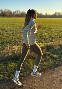Tenue de course comprenant une veste gris clair, des leggings vert olive et des chaussures de sport légères. L'arrière-plan montre un champ vert et des arbres.