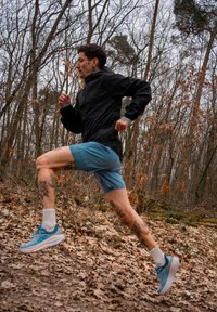 Corredor de casaco preto e calções azuis, com tatuagens visíveis nas pernas, no ar sobre um caminho de floresta coberto de folhas, usando sapatos de corrida azuis.