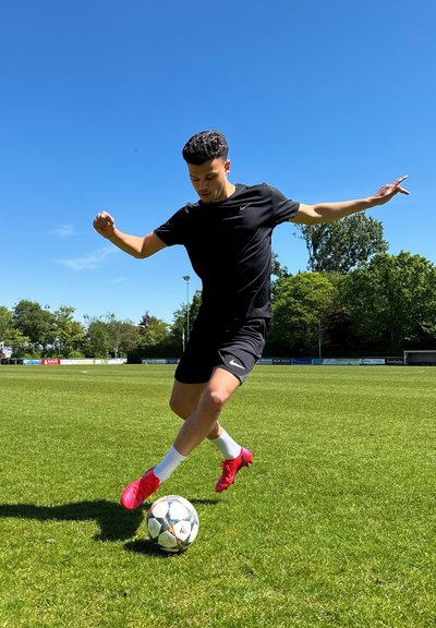 Atleta con ropa negra de Nike, realizando una patada en un campo de hierba, usando zapatos de fútbol de color rosa brillante. Se puede ver un balón de fútbol a sus pies.