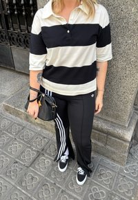 Person klädd i en svart- och vitrandig polotröja, svarta Adidas träningsbyxor med vita ränder, svarta sneakers och håller en svart handväska.