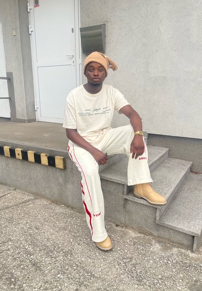 Gorro beige, camiseta crema con estampado, pantalones blancos con rayas rojas, botas beige, sentado en escalones grises al lado de la entrada.