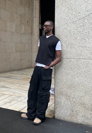 Top de punto negro sin mangas sobre una camiseta blanca, combinado con pantalones cargo negros con múltiples bolsillos y zapatos beige. Fondo de pared de piedra.