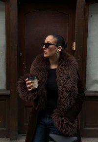 Casaco de pelo sintético castanho com uma textura peluda, blusa preta, jeans azuis e óculos de sol pretos oversized. Segurando uma chávena de café, perto de uma porta de madeira.