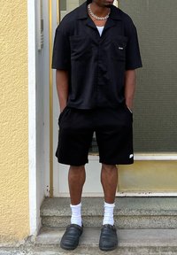 Musträne lühikese varrukaga nööbiga särk, millel on kaks taskut, sobivad mustad tekstuuriga lühikesed püksid, valged sokid ja mustad libisemiskindlad jalanõud.