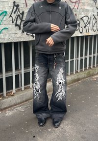 Grey zip-up sweatshirt with a rounded design, teamed with loose black jeans featuring large distressed patches. Black shoes complete the outfit.