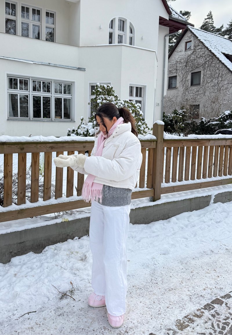 Donna con abiti invernali bianchi e sciarpa rosa che si mette i guanti accanto a una recinzione di legno fuori da case innevate.