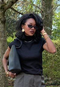 Top preto de manga curta, com gola alta, combinado com calças cinzentas claras. Os acessórios incluem brincos grandes dourados, uma pulseira e uma bolsa preta oversized.